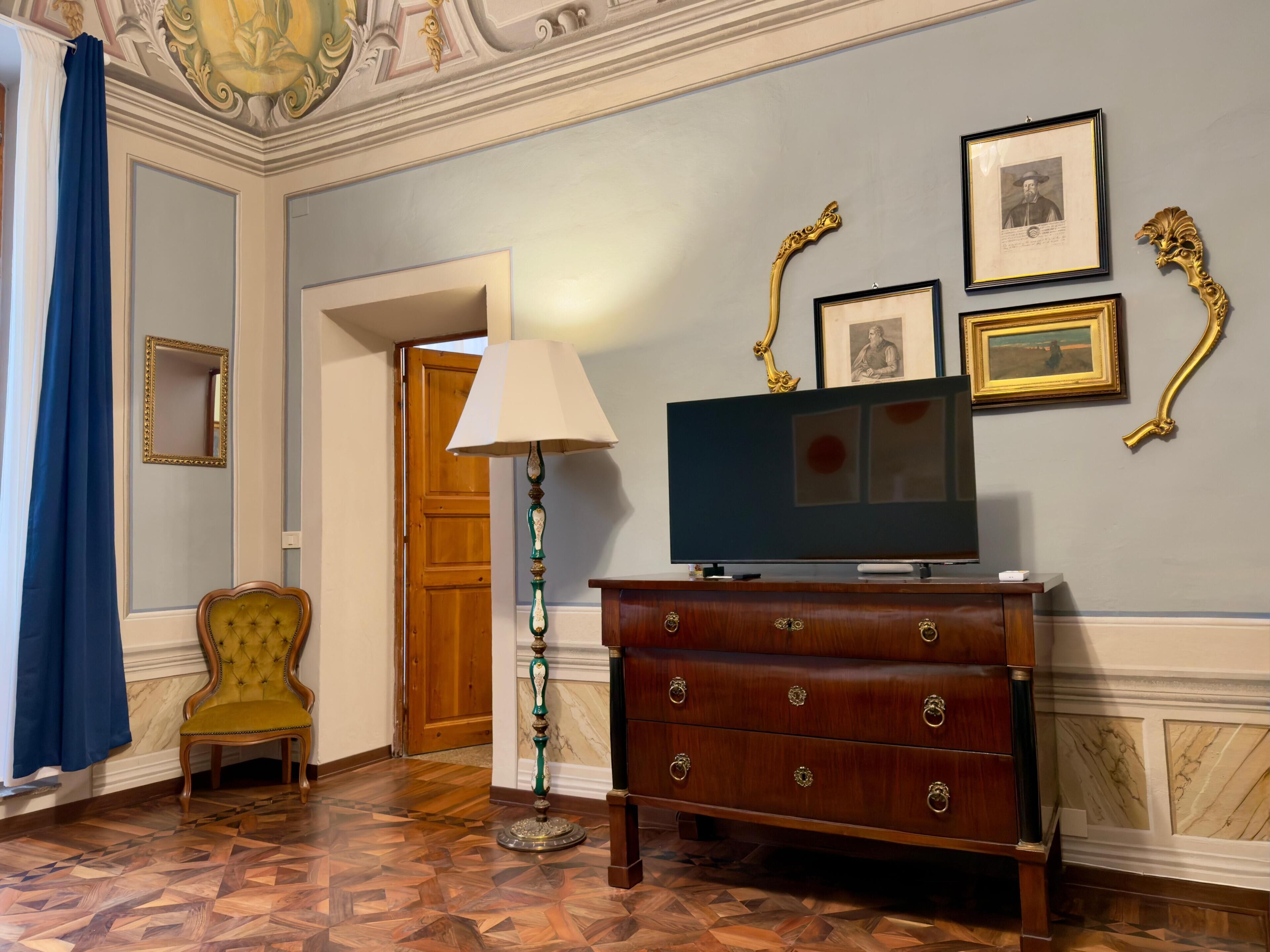 Une pièce avec un plancher en bois, un téléviseur posé sur une commode en bois, une lampe, une chaise et des tableaux encadrés accrochés au mur.