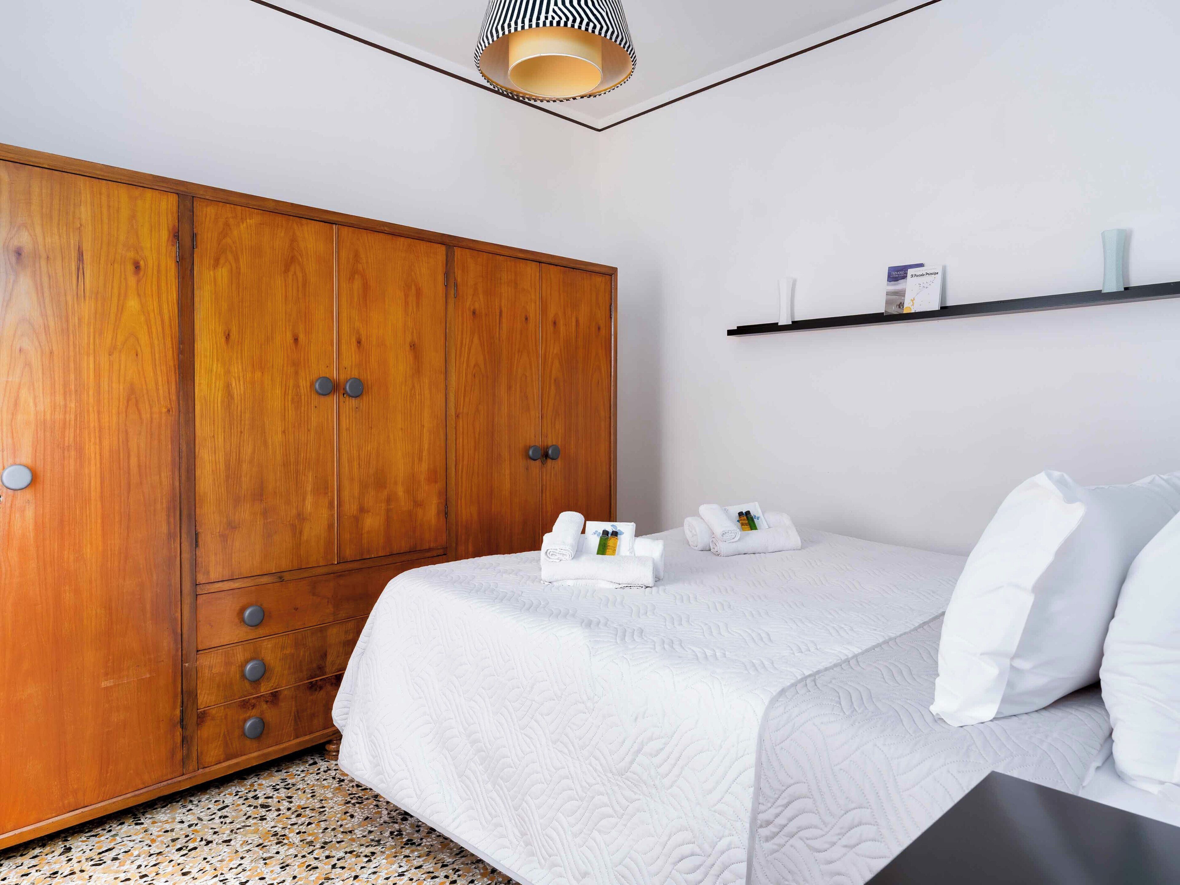 Un dormitorio con un armario de madera, una cama con ropa de cama blanca y un estante pequeño con artículos de aseo.