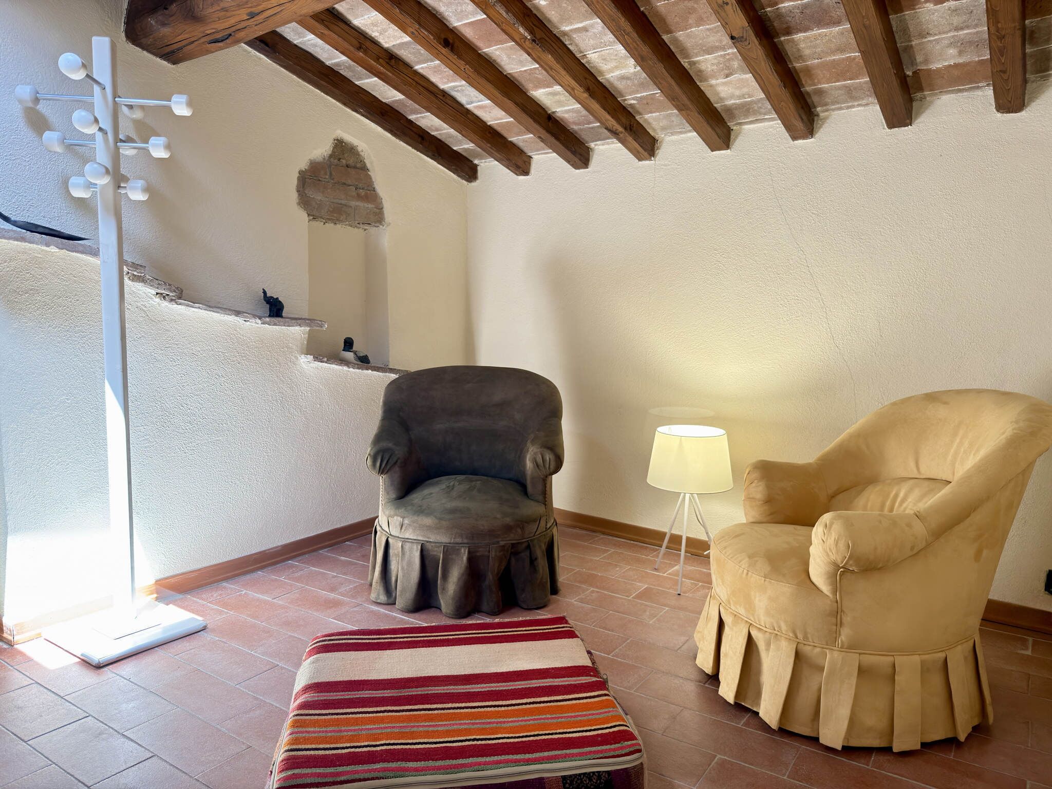 Una habitación con suelo de baldosas, techo de madera, un sillón gris, un sillón beige, una mesita con una lámpara y una alfombra a rayas.