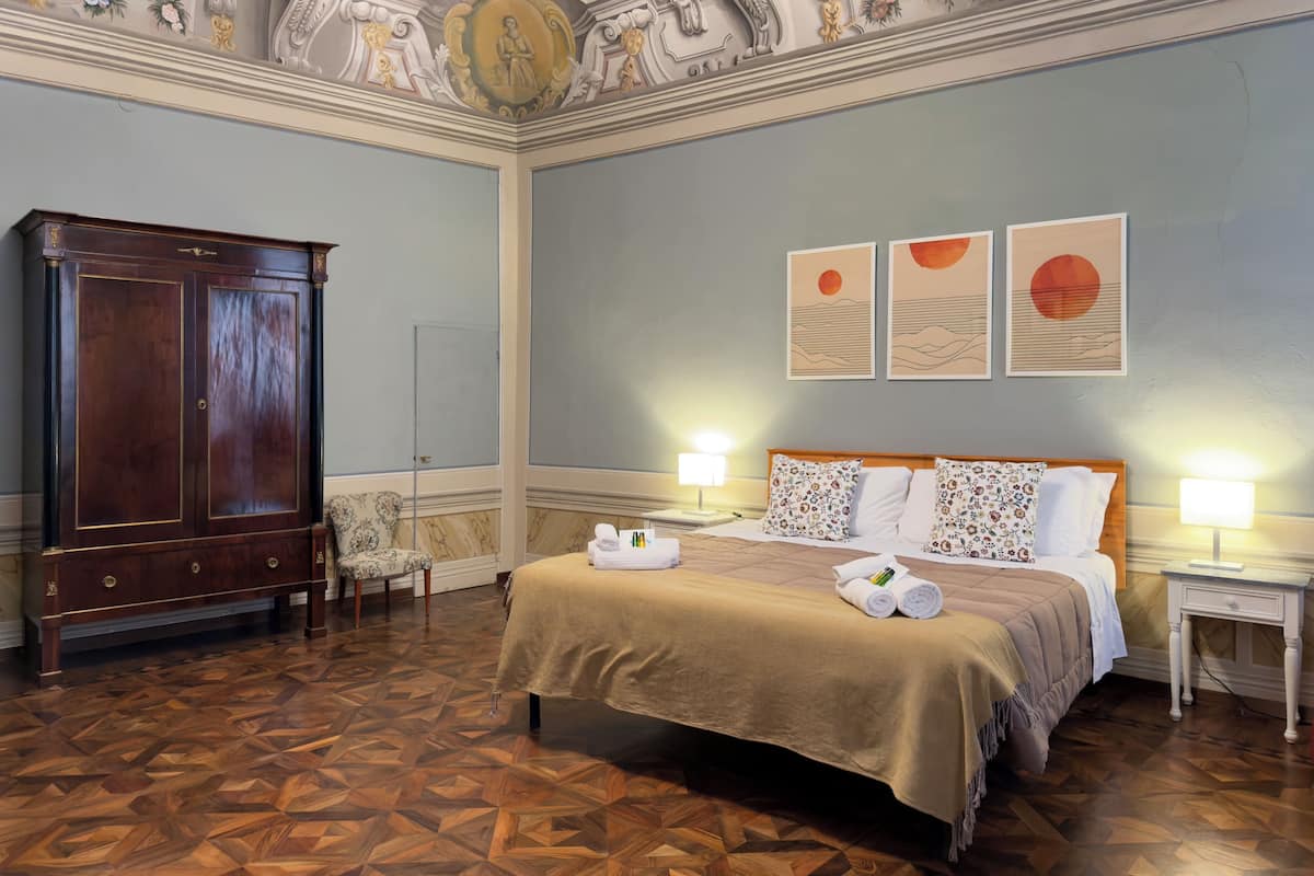 A room with a bed, a wooden wardrobe, a chair, and a nightstand with a lamp.