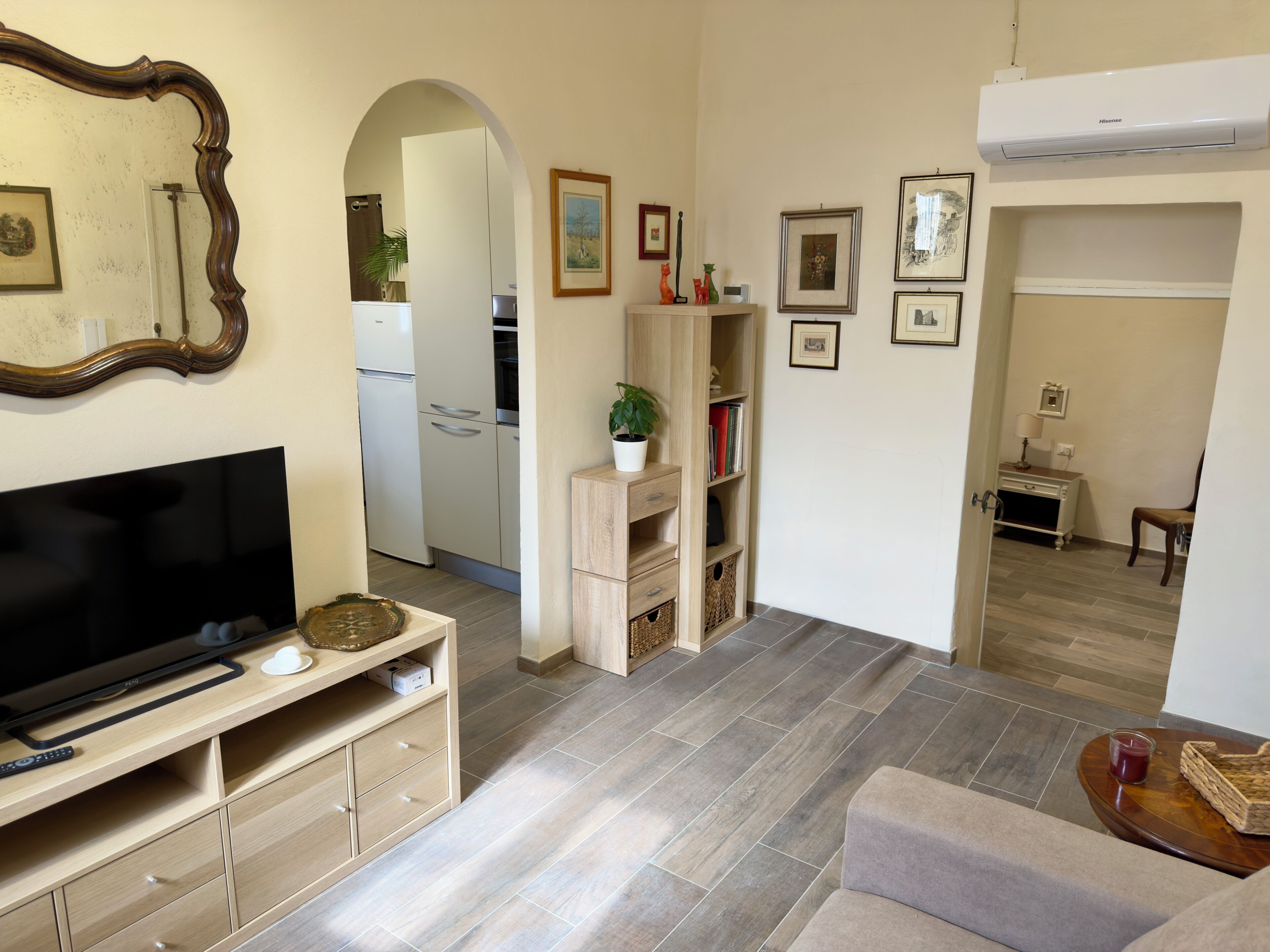 Ein Wohnzimmer mit Flachbildfernseher, einer Holz-Unterhaltungsecke und einer Couch. Im Hintergrund ist eine Küche mit Kühlschrank und Schränken zu sehen.