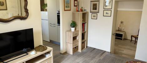 Ein Wohnzimmer mit Flachbildfernseher, einer Holz-Unterhaltungsecke und einer Couch. Im Hintergrund ist eine Küche mit Kühlschrank und Schränken zu sehen.