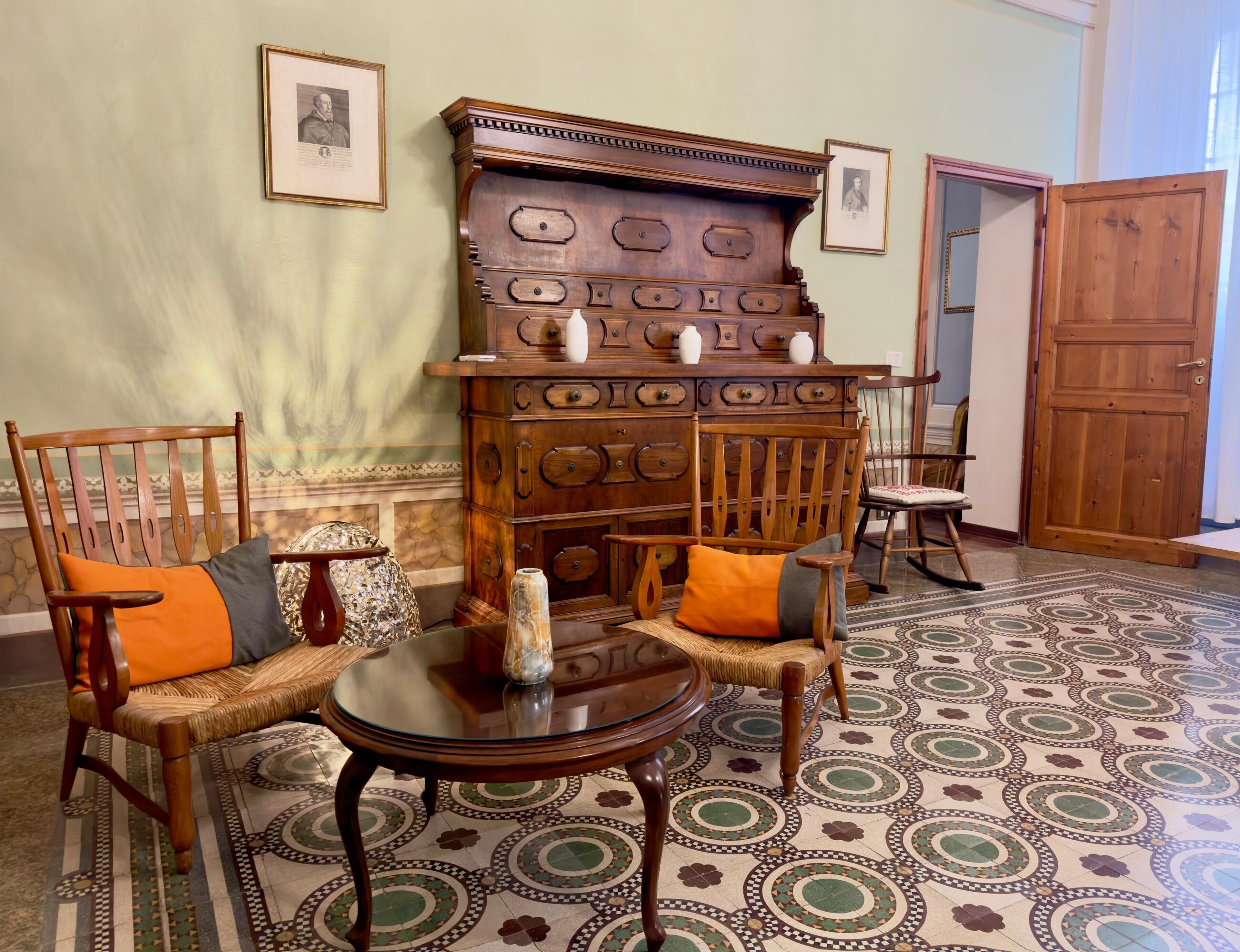 A room with a large wooden cabinet, two wooden chairs with cushions, a small round table, patterned floor tiles, and a wooden door.