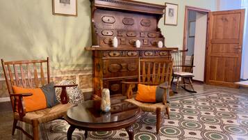 A room with a large wooden cabinet, two wooden chairs with cushions, a small round table, patterned floor tiles, and a wooden door.