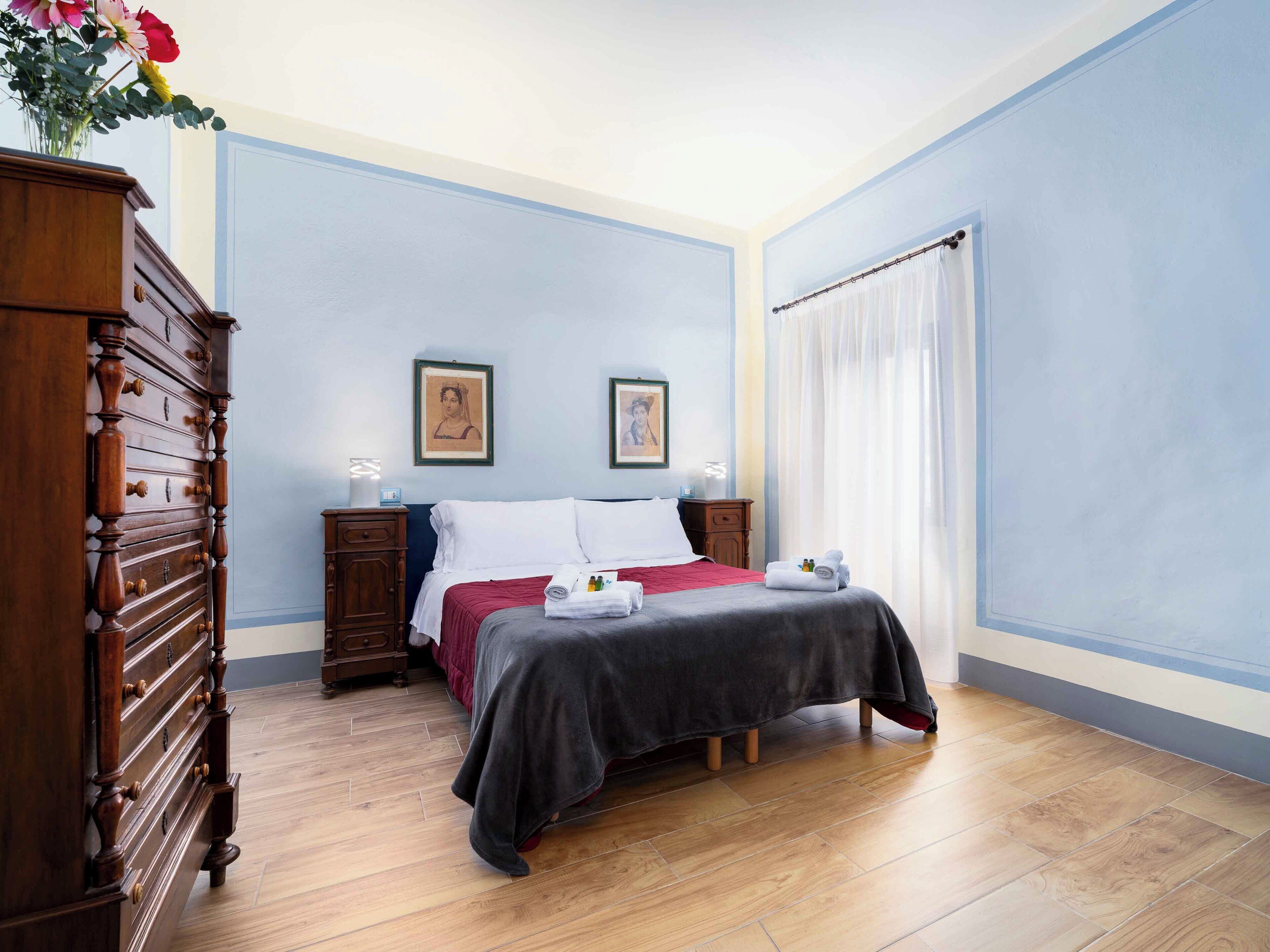 Een hotelkamer met een houten dressoir, een bed met een rode sprei, twee ingelijste foto's aan de muur en een raam met witte gordijnen.