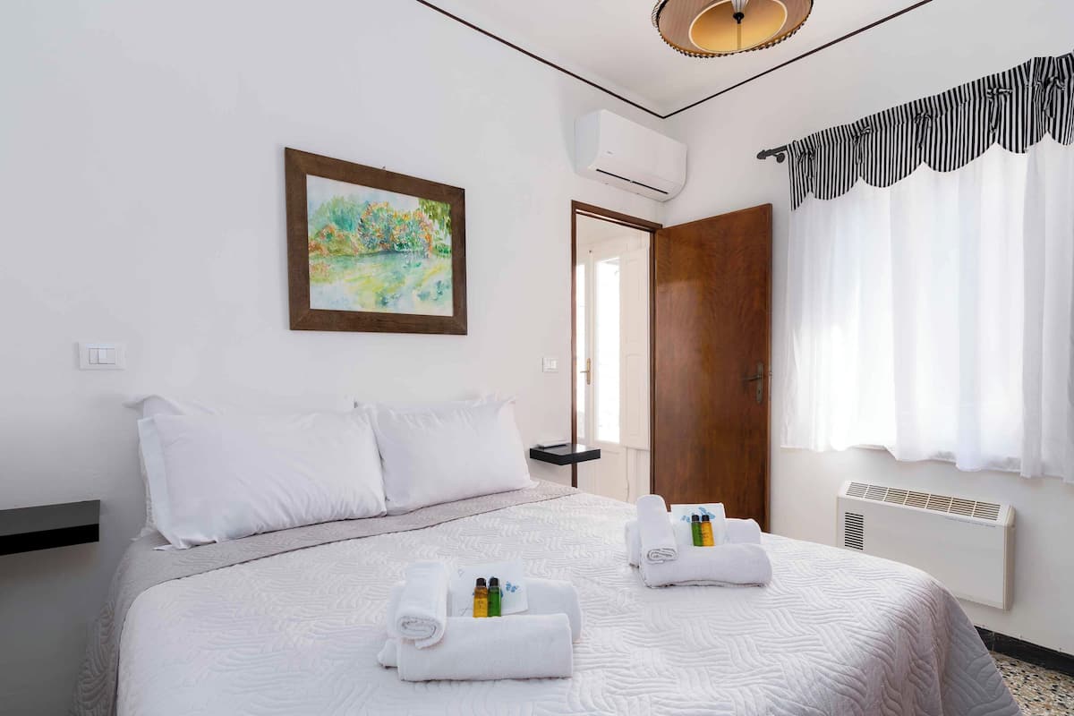 A hotel room with a bed, a painting, an air conditioning unit, a window with curtains, and a door.