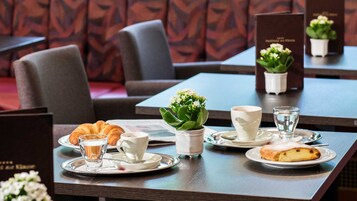 Una mesa con platos, un croissant, un vaso de agua, una taza y una pequeña planta en maceta.