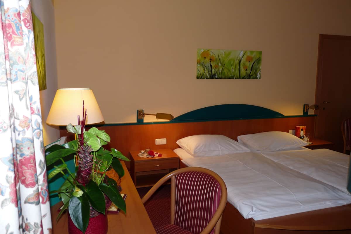 A hotel room with a bed, a nightstand, a lamp, a chair, a potted plant, and a painting on the wall.