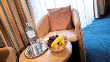 A hotel room with a round wooden table, a wicker fruit basket with grapes, a banana, and an apple, a bottle of water, and a glass on the table.