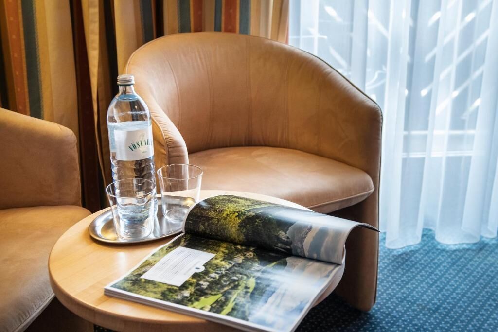 Habitación de hotel con un sillón de cuero, una mesita redonda con una revista, una botella de agua y vasos.
