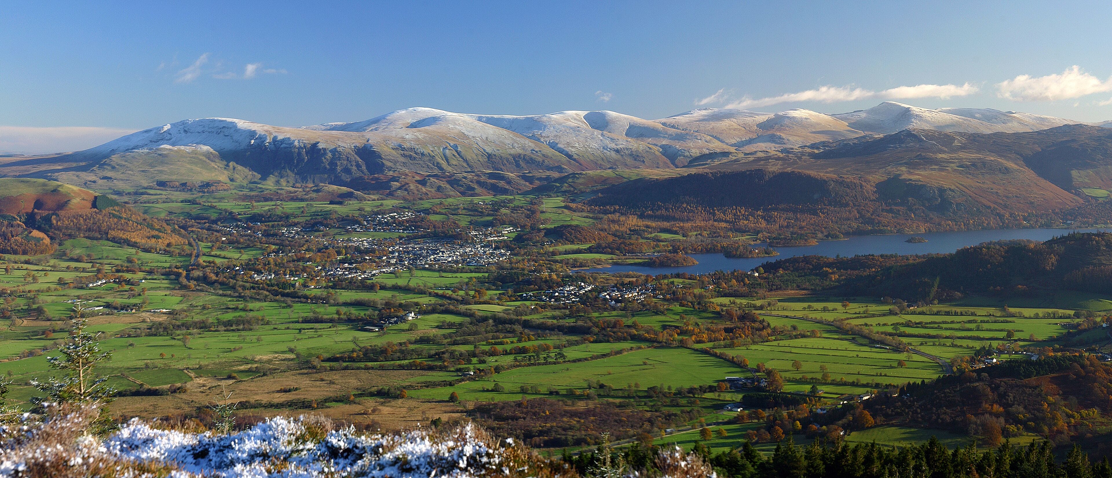 Una vista panorámica de un valle con un lago, un pueblo pequeño y montañas nevadas al fondo.