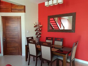 A dining area with a wooden table and chairs, a vase with flowers, and a red wall with a mirror reflecting a staircase.