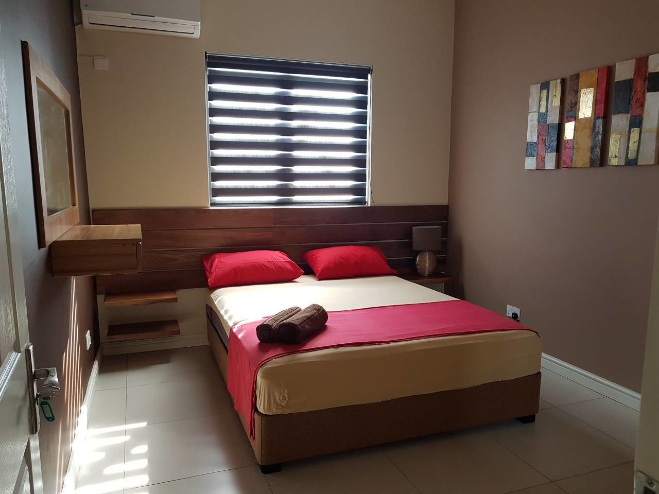 Un dormitorio con cama, ventana con persianas, mesita de noche y una obra de arte en la pared.