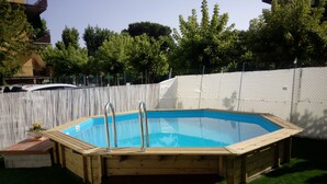 Una piscina elevada de madera con escaleras de metal, rodeada por una valla blanca y césped verde.