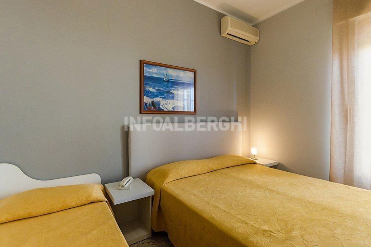 Quarto de hotel com duas camas, mesa de cabeceira com telefone, pintura de paisagem marítima e unidade de ar condicionado.