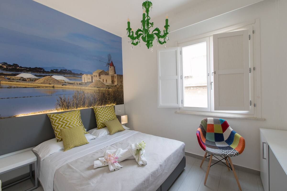 A bedroom with a bed, a colorful chair, a chandelier, and a large painting of a landscape.