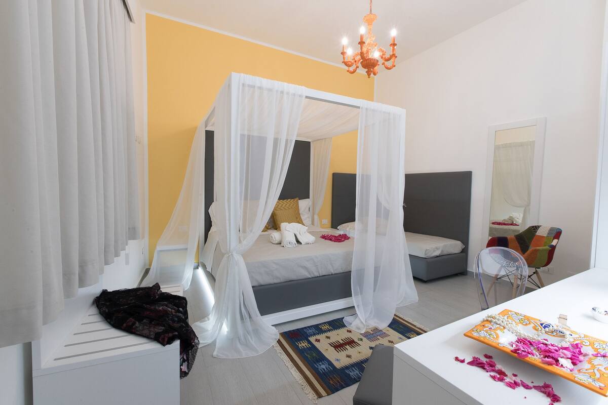 A bedroom with a four-poster bed, a bedside table, a chair, and a desk with a mirror.