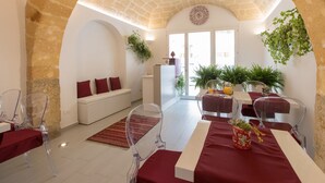 A dining area with a stone ceiling, white walls, and red accents. There are clear chairs, a white sofa with red cushions, and a table with a red tablecloth. - B&B Marsala (Marsala)