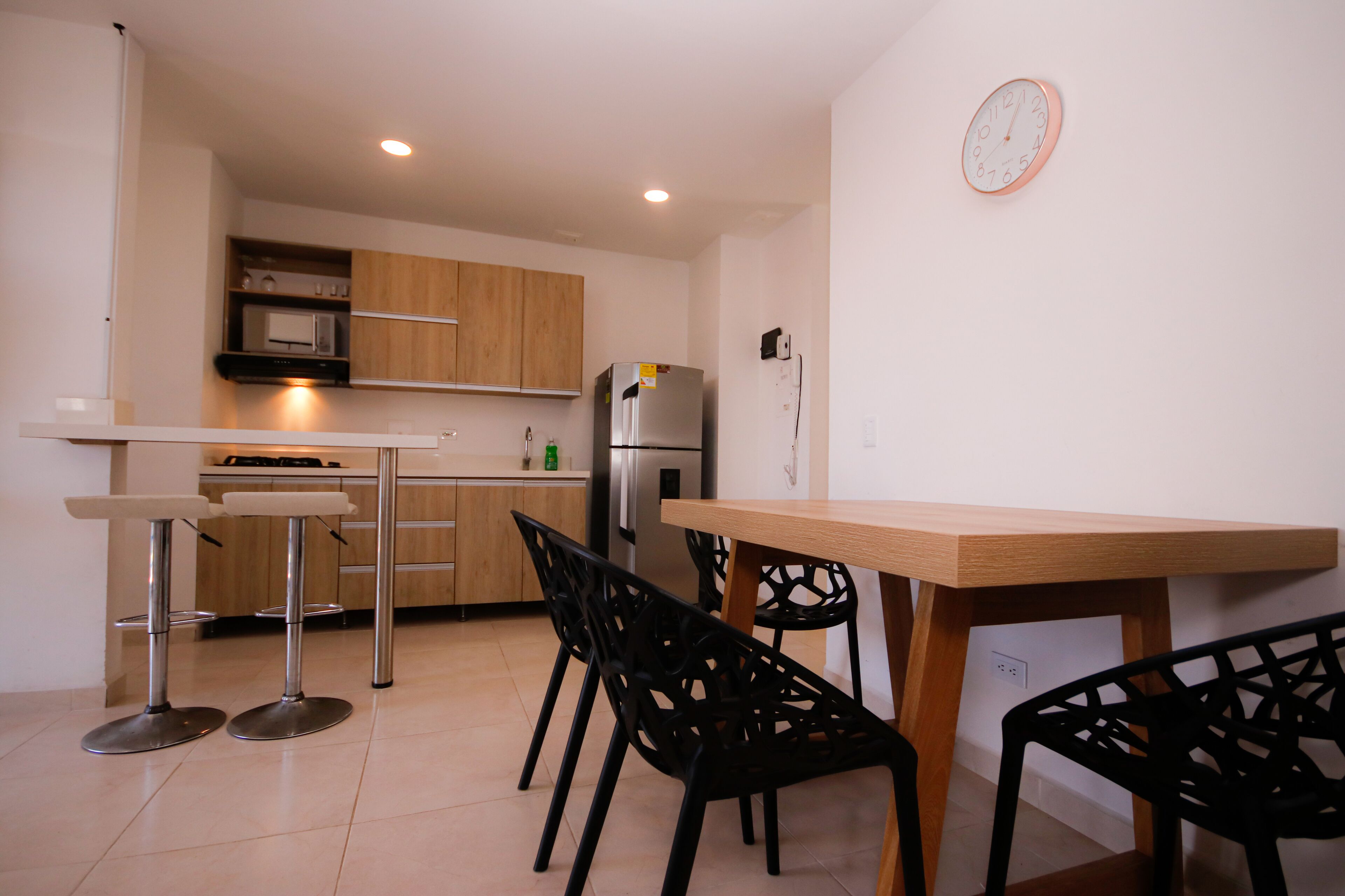 A modern kitchen with wooden cabinets, a stainless steel refrigerator, a microwave, and a wooden dining table with black chairs.