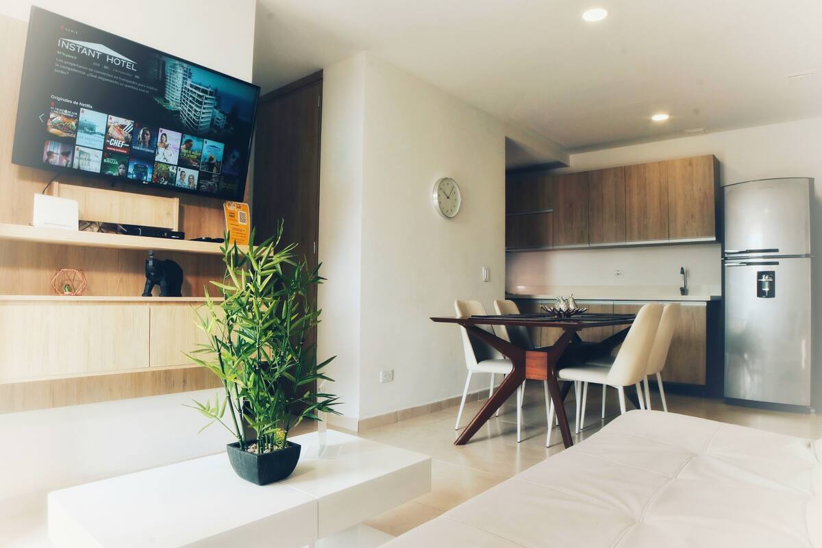 Un salon moderne doté d’un téléviseur à écran plat affichant l’interface de Netflix, d’un coin repas avec une table et des chaises en bois, et d’une cuisine avec des meubles en bois et un réfrigérateur en acier inoxydable.