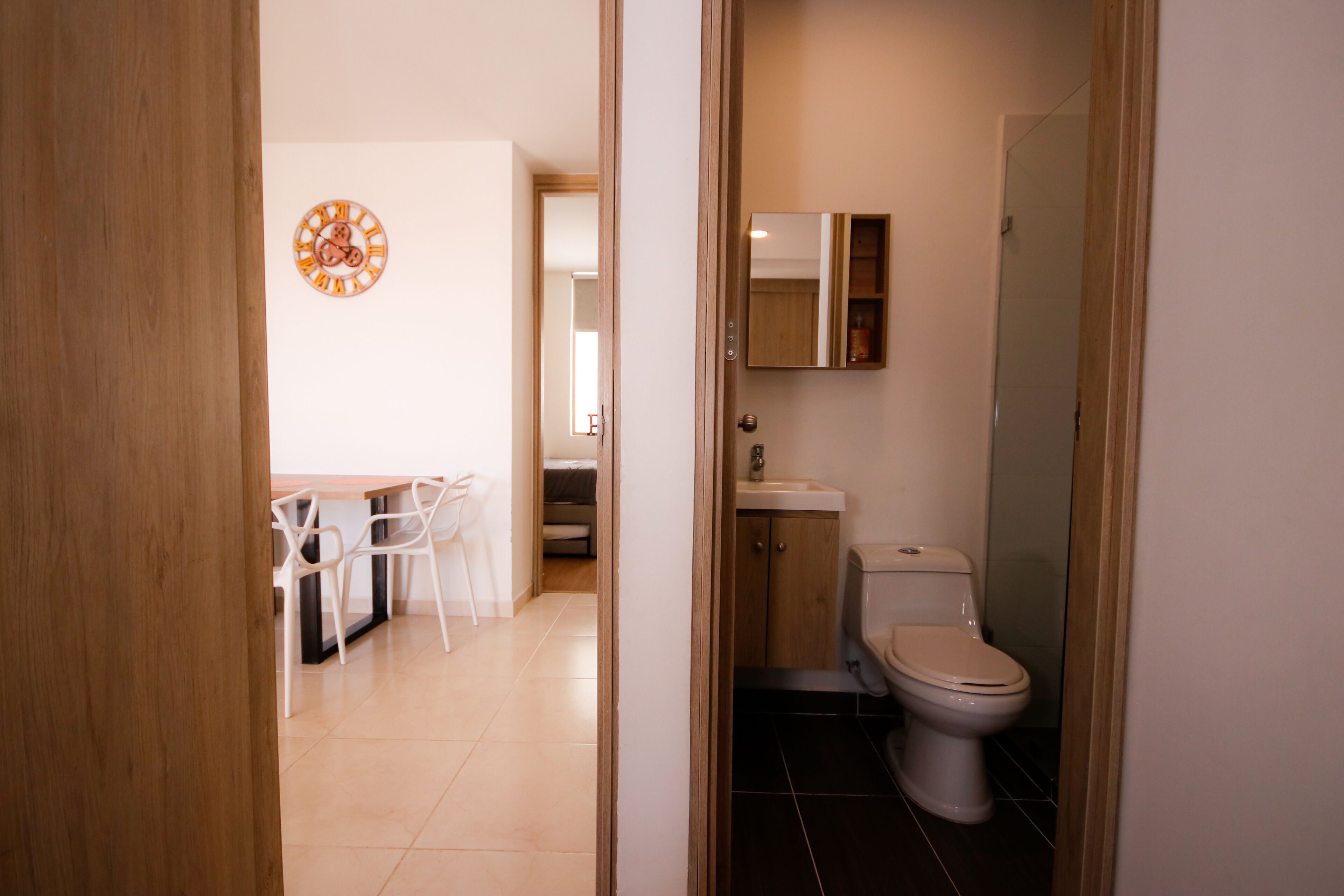 A modern bathroom with a toilet, sink, and mirror. There is a dining area with chairs and a table visible through an open door.