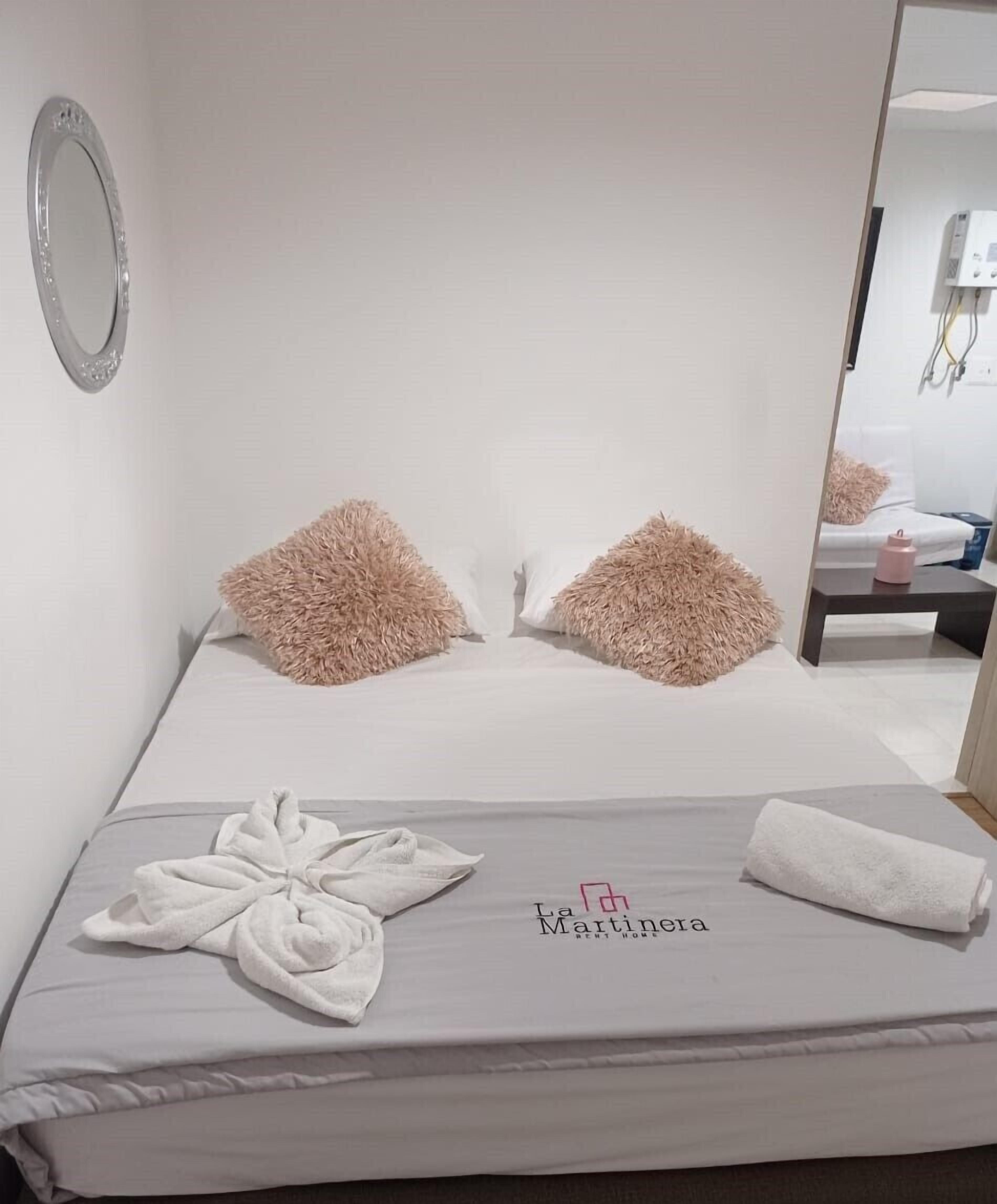 A neatly made bed with a towel and a decorative pillow, a mirror on the wall, and a small bedside table with a pink object.