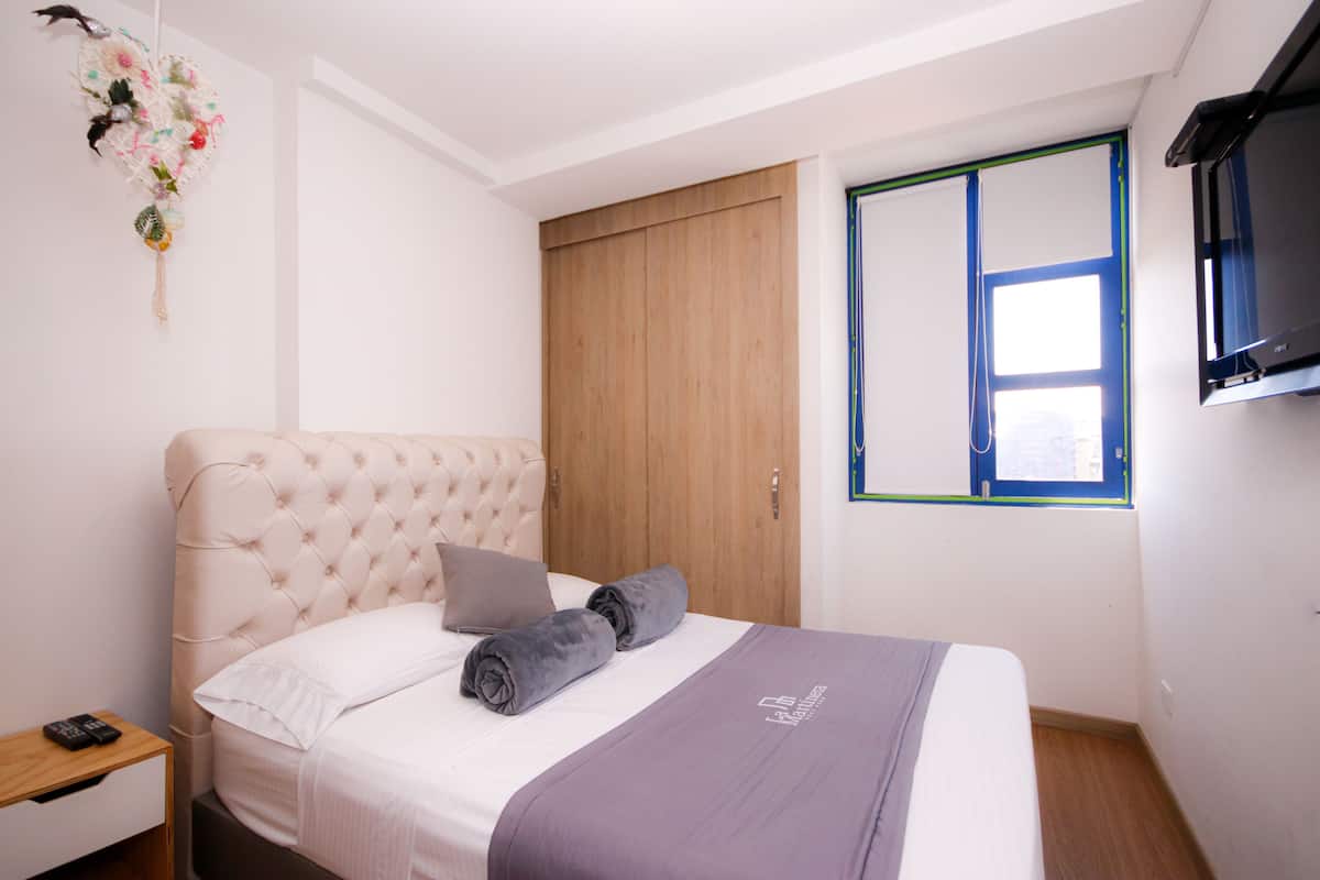 Une chambre à coucher bien aménagée, avec une tête de lit capitonnée, un lit recouvert d’une literie blanche et violette, une table de chevet en bois et un téléviseur fixé au mur.