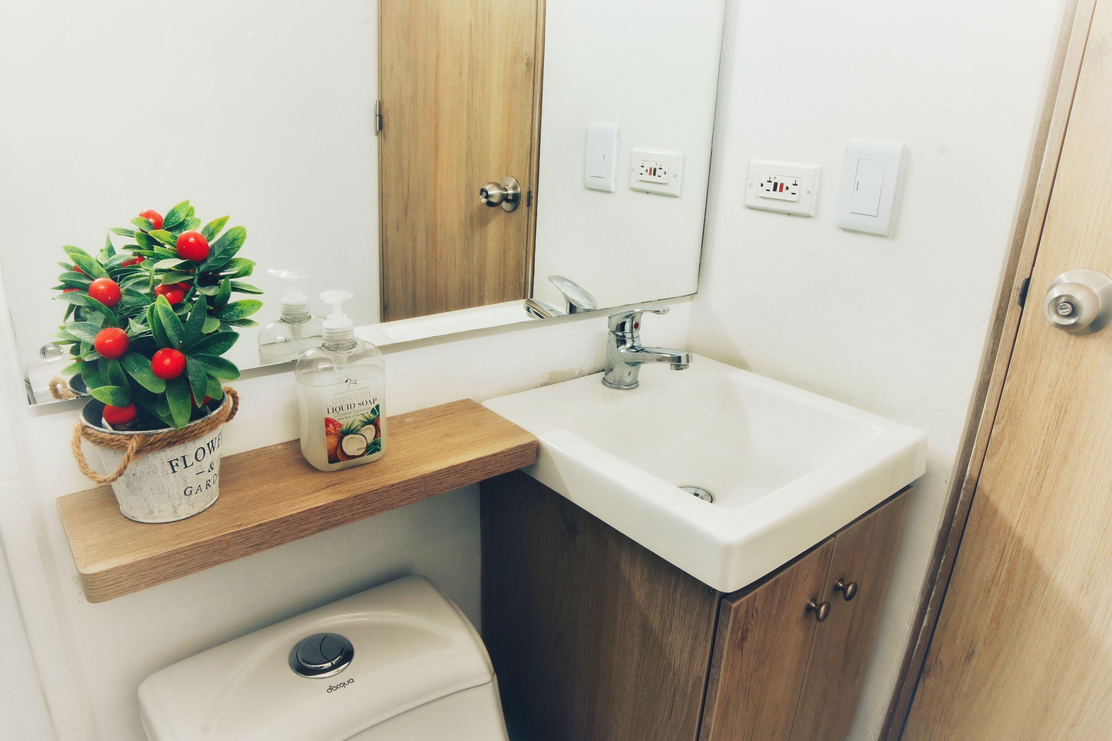 A bathroom with a wooden vanity, a white sink, a mirror, a potted plant, and a toilet.
