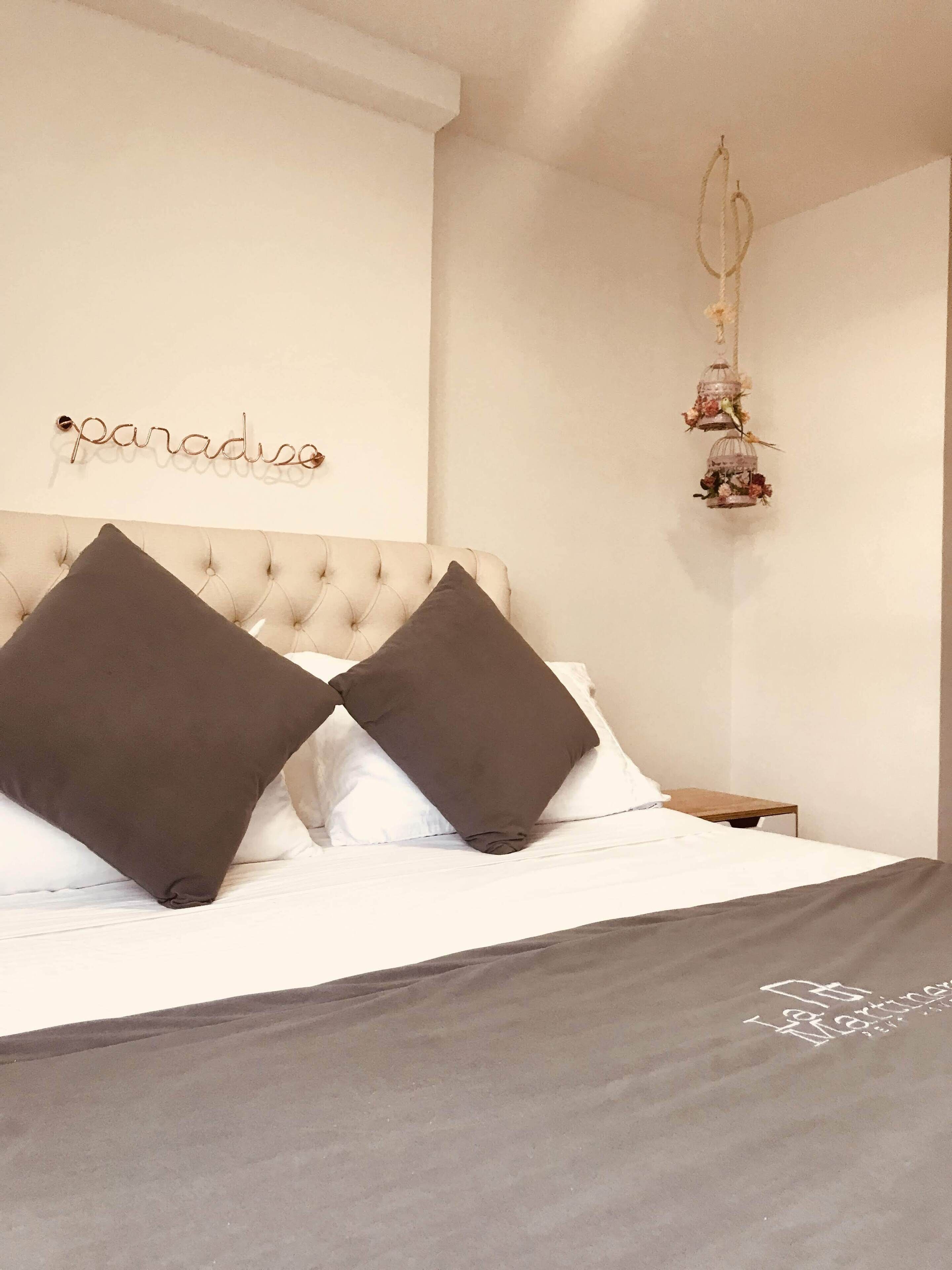 A bedroom with a tufted headboard, two dark pillows, a white bedspread, a small bedside table, and a decorative birdcage with flowers hanging from the ceiling.