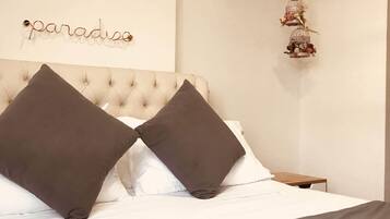 A bedroom with a tufted headboard, two dark pillows, a white bedspread, a small bedside table, and a decorative birdcage with flowers hanging from the ceiling.