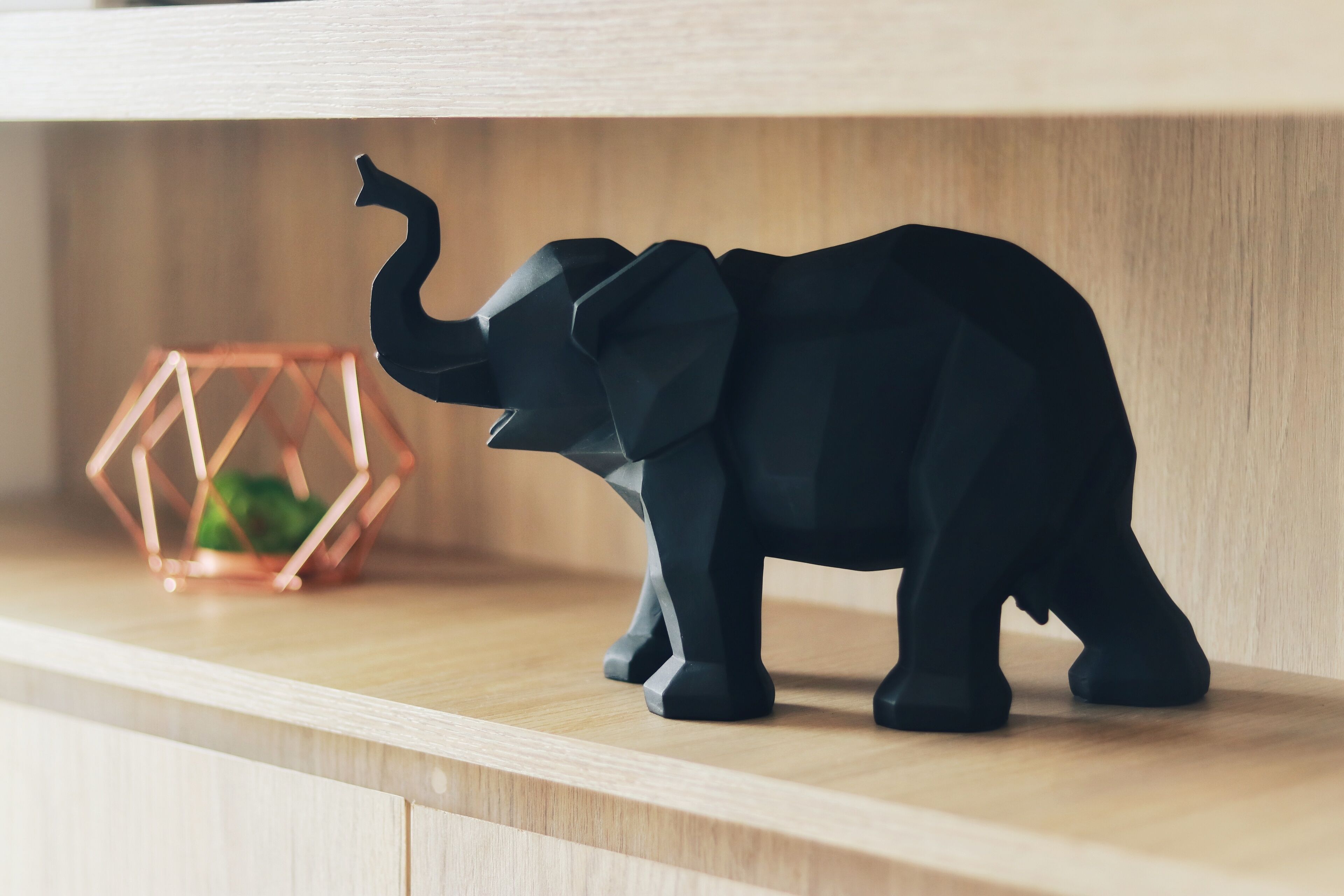 Una figurita de elefante negro sobre una repisa de madera, con un objeto de color cobre y diseño geométrico al fondo.
