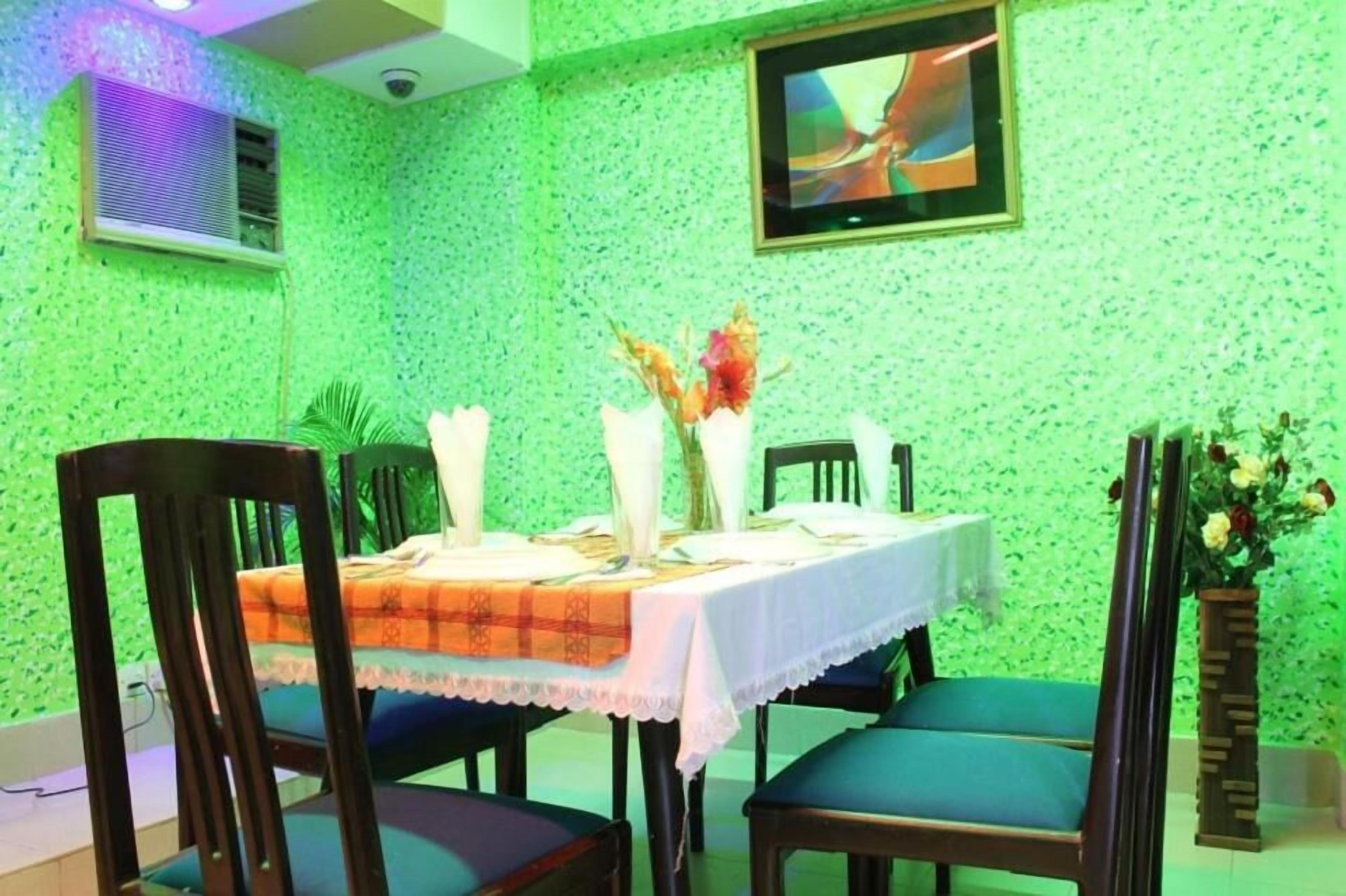 A dining area with a green textured wall, a television mounted above the table, and a vase with flowers on the table.