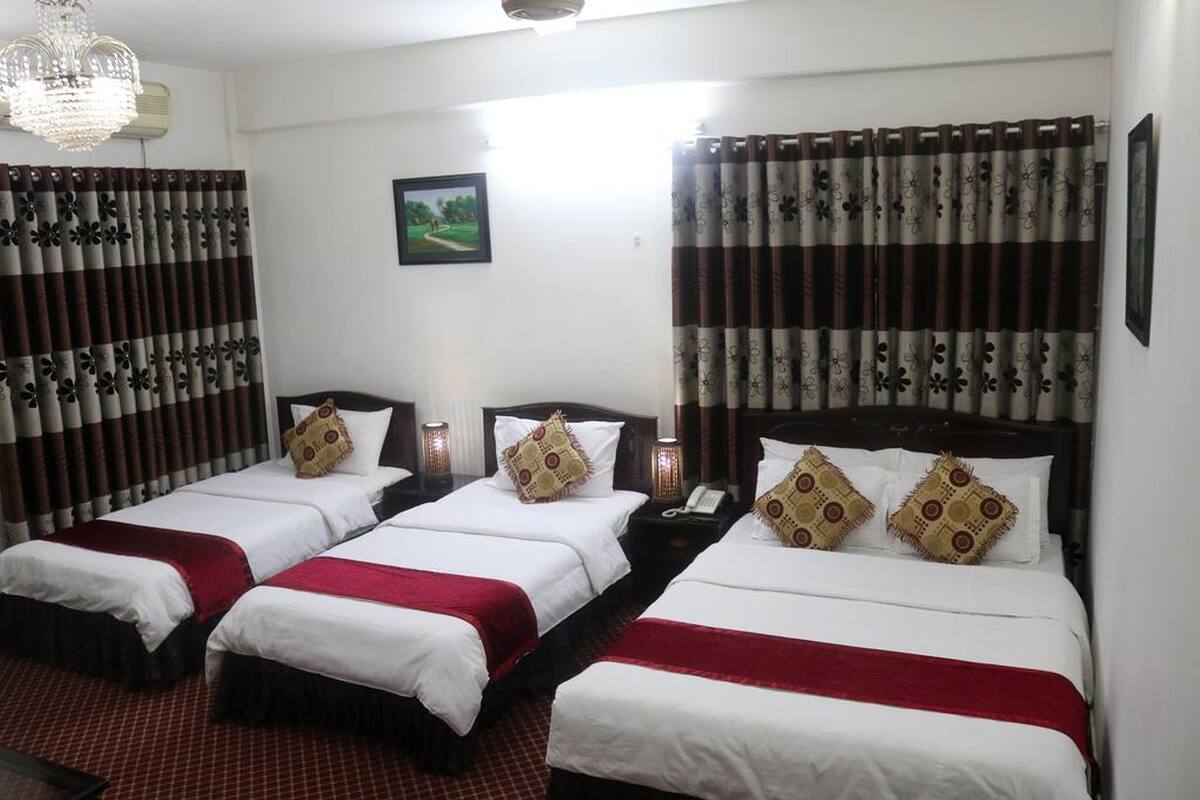A hotel room with three single beds, each with a red and white bedspread, and a large bed with a red and white bedspread. There are two nightstands with lamps, a chandelier, and a framed picture on the wall.