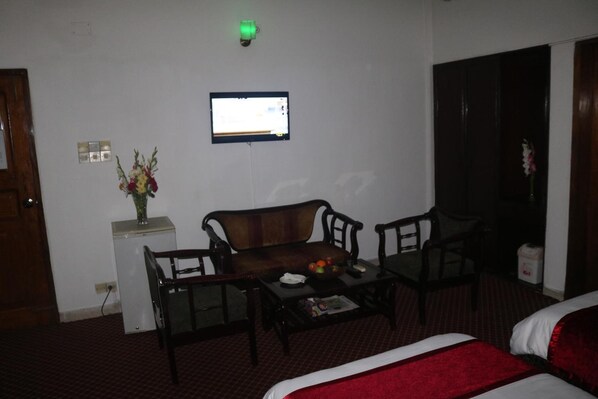 A hotel room with a bed, a desk, a chair, a small table with a vase of flowers, and a television on the wall. - Hotel De Meridian (Dhaka)