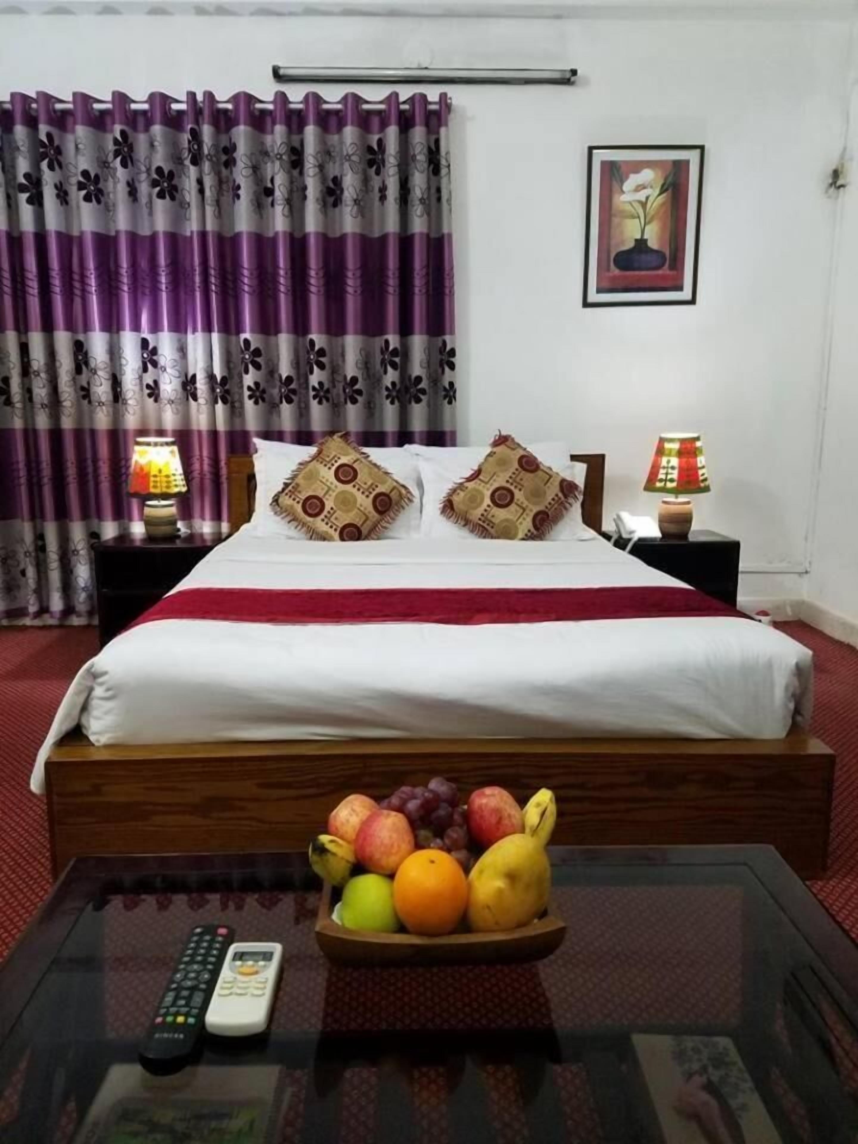 A neatly made bed with a red blanket, two decorative pillows, a fruit bowl on the bedside table, a remote control, and a lamp on a nightstand.