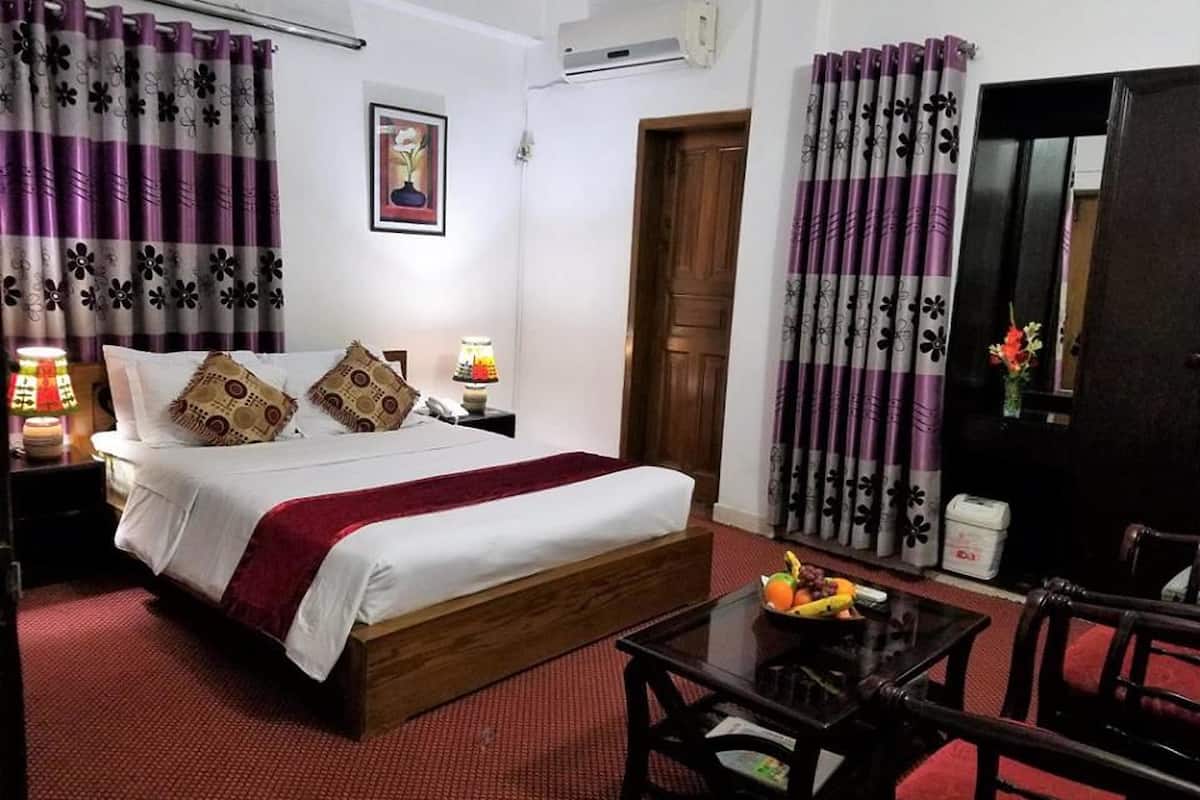 A hotel room with a wooden bed, red carpet, purple curtains, a bedside table with a lamp, a small table with fruit, and a chair.