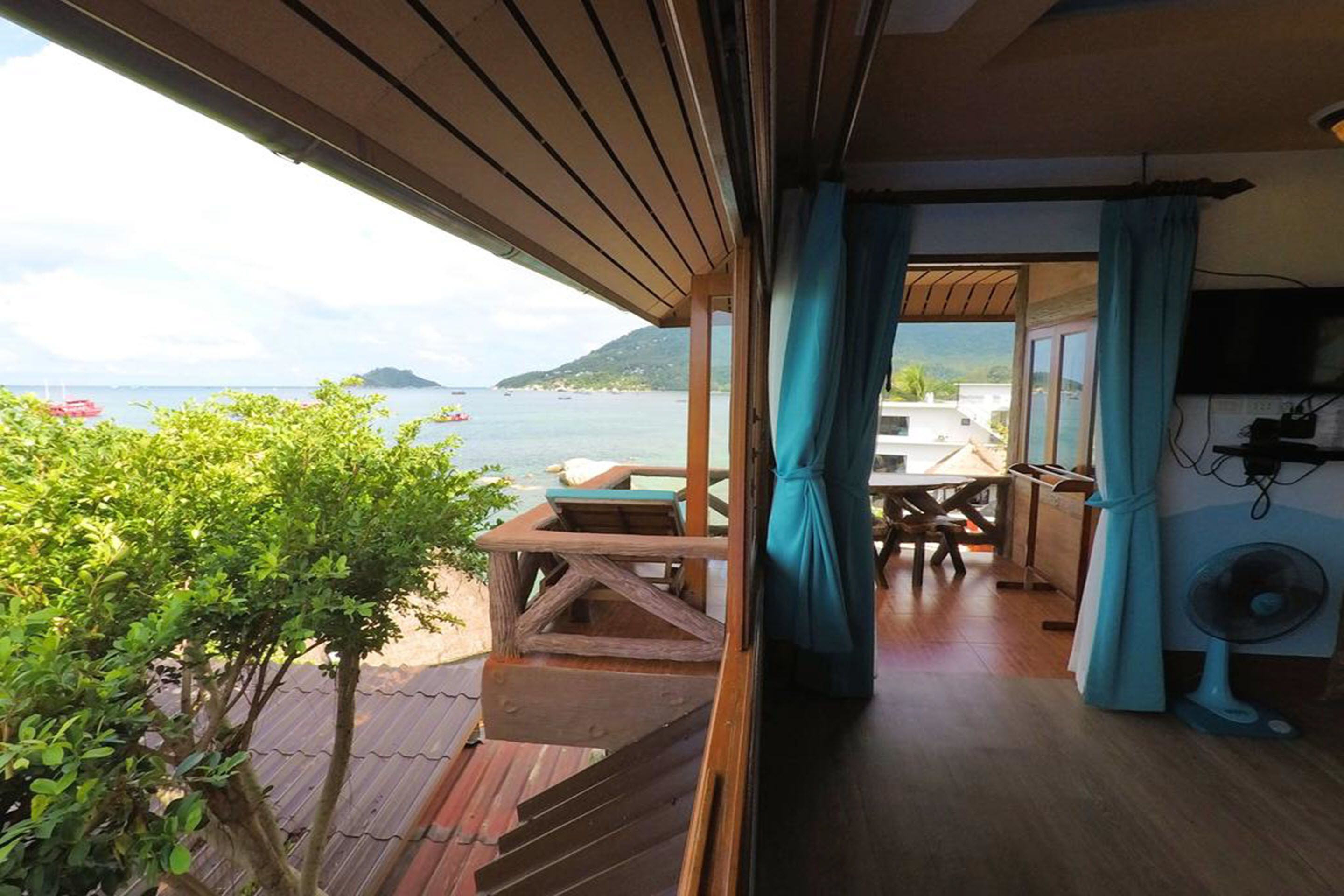 Eine Holzterrasse mit Tisch und Stühlen, einem Ventilator und Blick auf das Meer und die Hügel.
