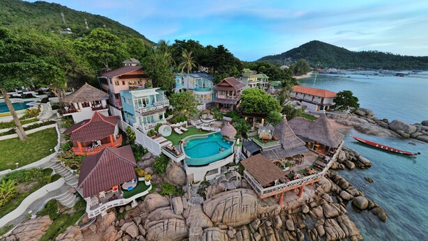 Ein Resort mit Swimmingpool, strohgedeckten Gebäuden und einer felsigen Küstenlinie.