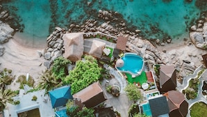 Luftaufnahme eines Resorts mit Schwimmbad, Strand und mehreren Gebäuden mit markanten Dächern.