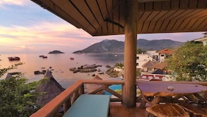Eine Holzterrasse mit Bank und Tisch mit Blick auf ein ruhiges Meer, Schiffe, eine felsige Küste und Gebäude im Hintergrund.