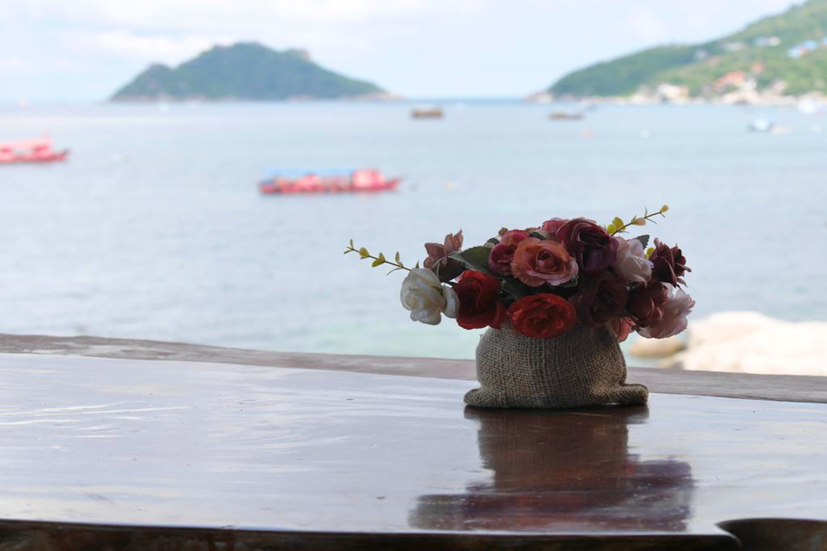 Un vase de fleurs est posé sur une table en bois, avec une vue sur la mer et des bateaux en arrière-plan.