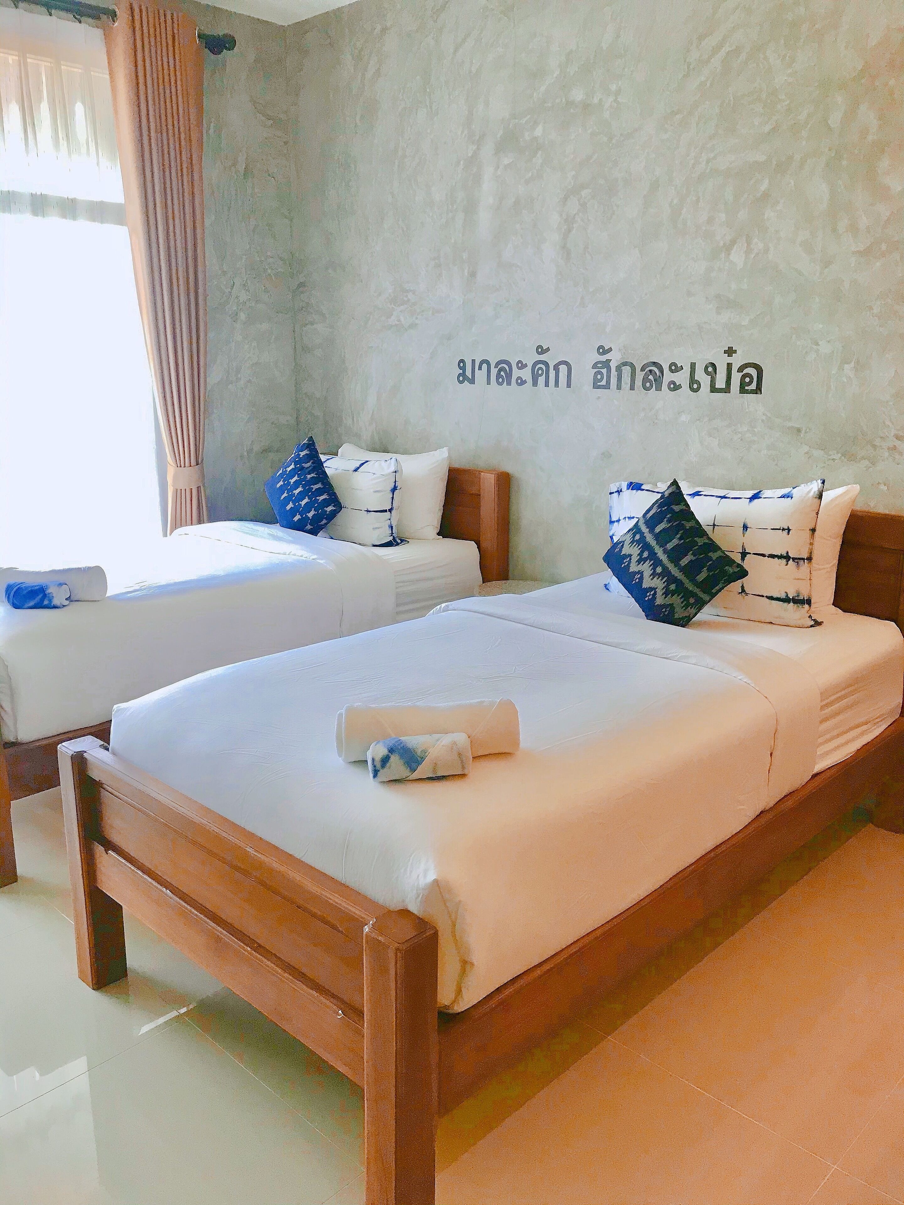 A hotel room with two beds, each with white linens and blue patterned pillows. The room has a concrete wall with Thai text and a window with curtains.
