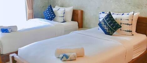 A hotel room with two beds, each with white linens and blue patterned pillows. The room has a concrete wall with Thai text and a window with curtains.