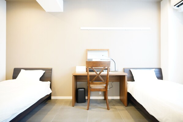 A hotel room with a bed, a desk with a chair, a trash bin, and a wall-mounted light fixture. - Sunshine Coast (Chatan)