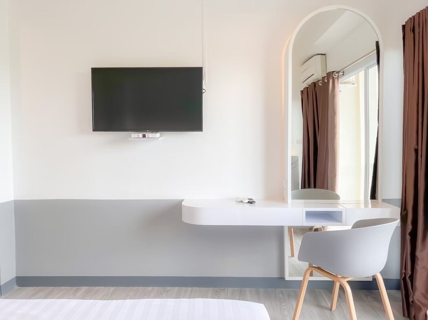 Una habitación de hotel moderna con un televisor de pantalla plana montado en la pared, un tocador blanco con espejo y una silla gris.