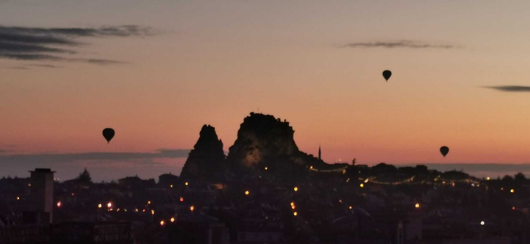 A cityscape at dusk with hot air balloons and a prominent rock formation.