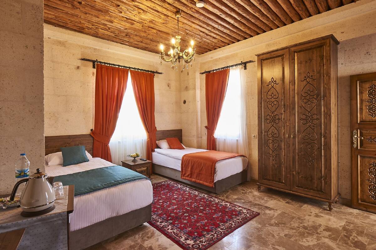 A hotel room with two beds, a wooden wardrobe, a chandelier, and a red patterned rug.