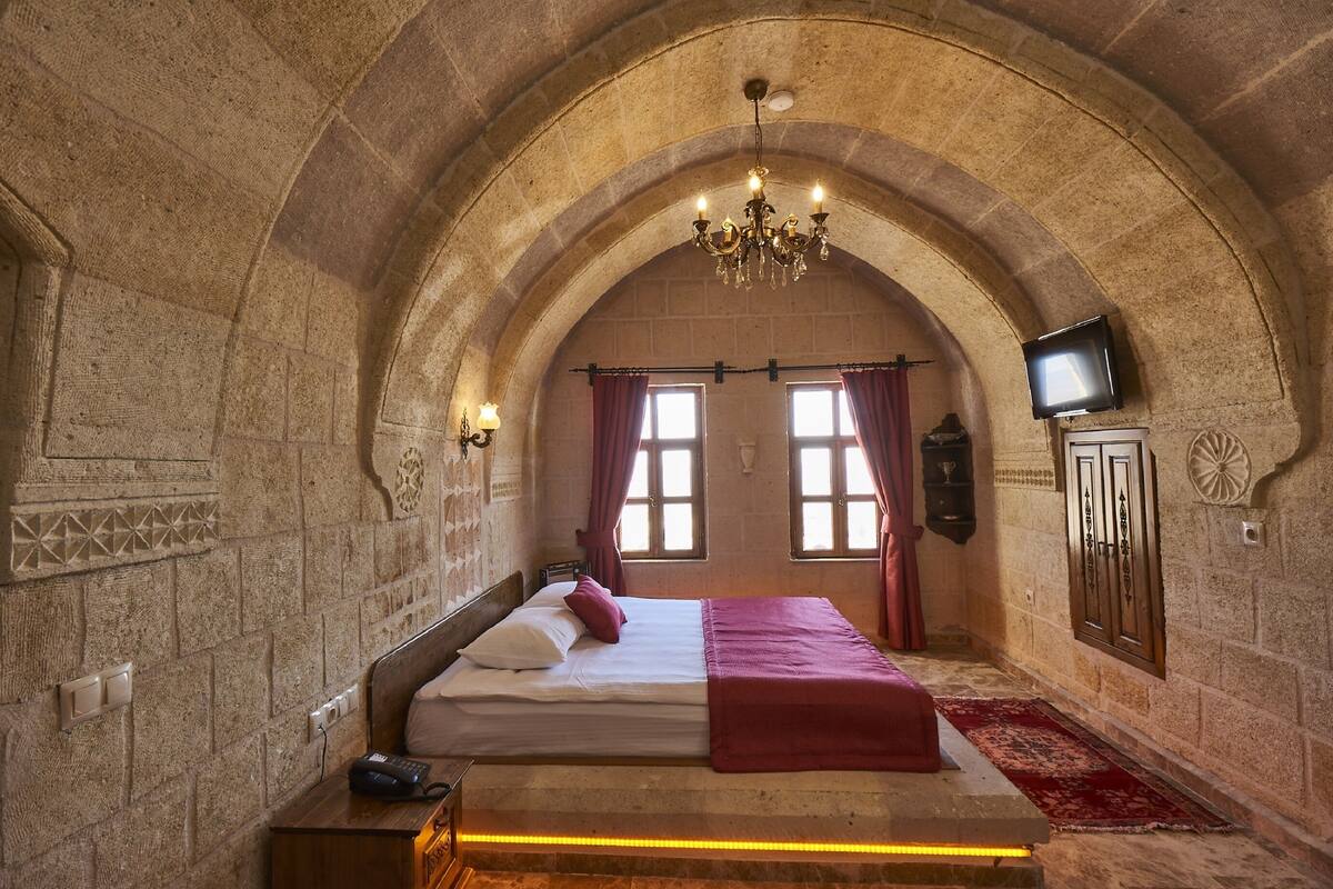 A room with a stone arch ceiling, a bed with a red blanket, a chandelier, a television on the wall, and a red carpet.