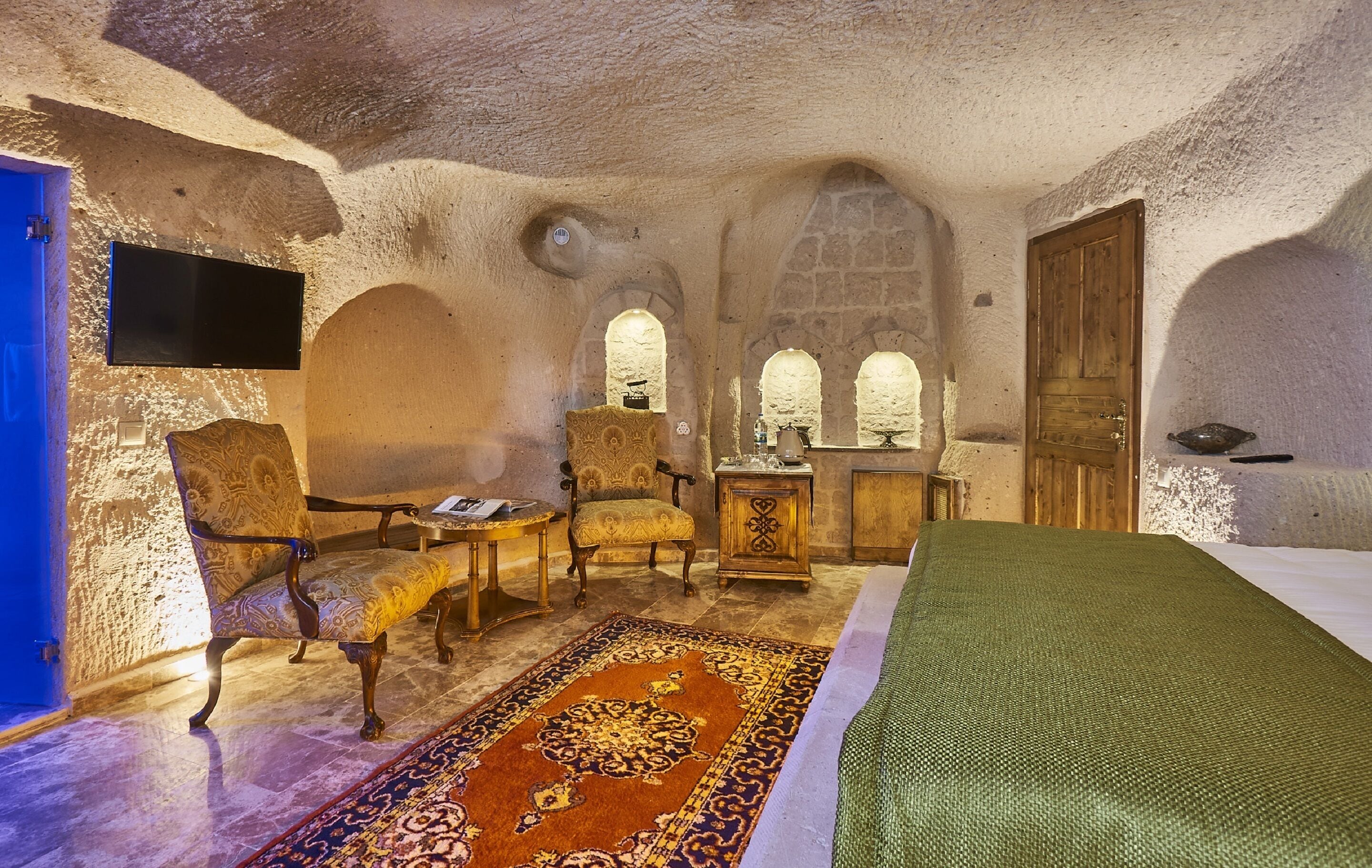 A room with a bed, two armchairs, a small table, and a television mounted on the wall.