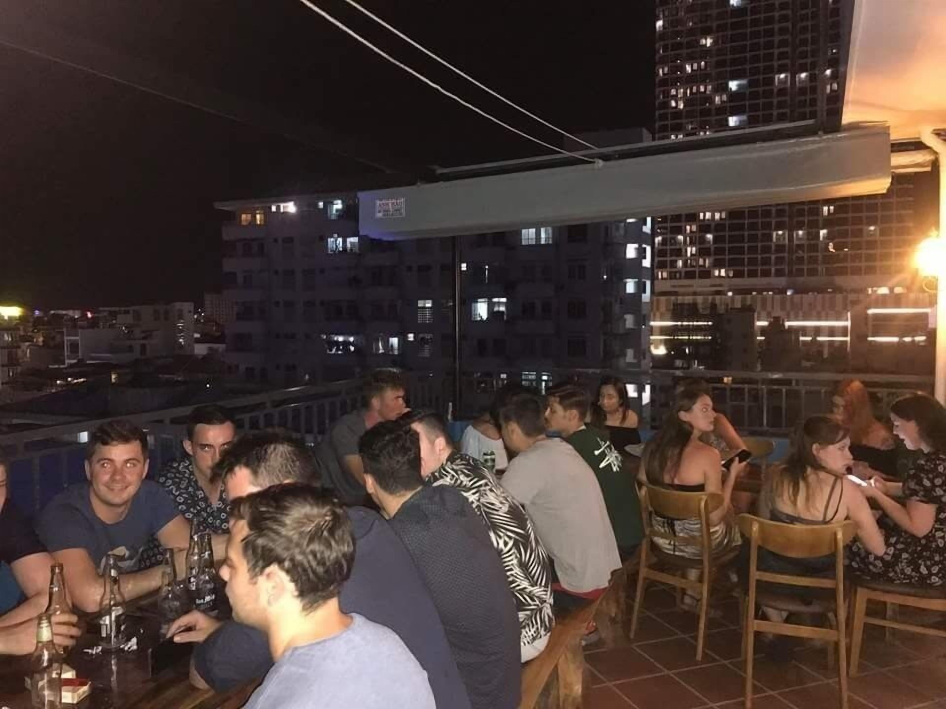 A rooftop bar with people seated at tables, overlooking a cityscape with illuminated buildings.